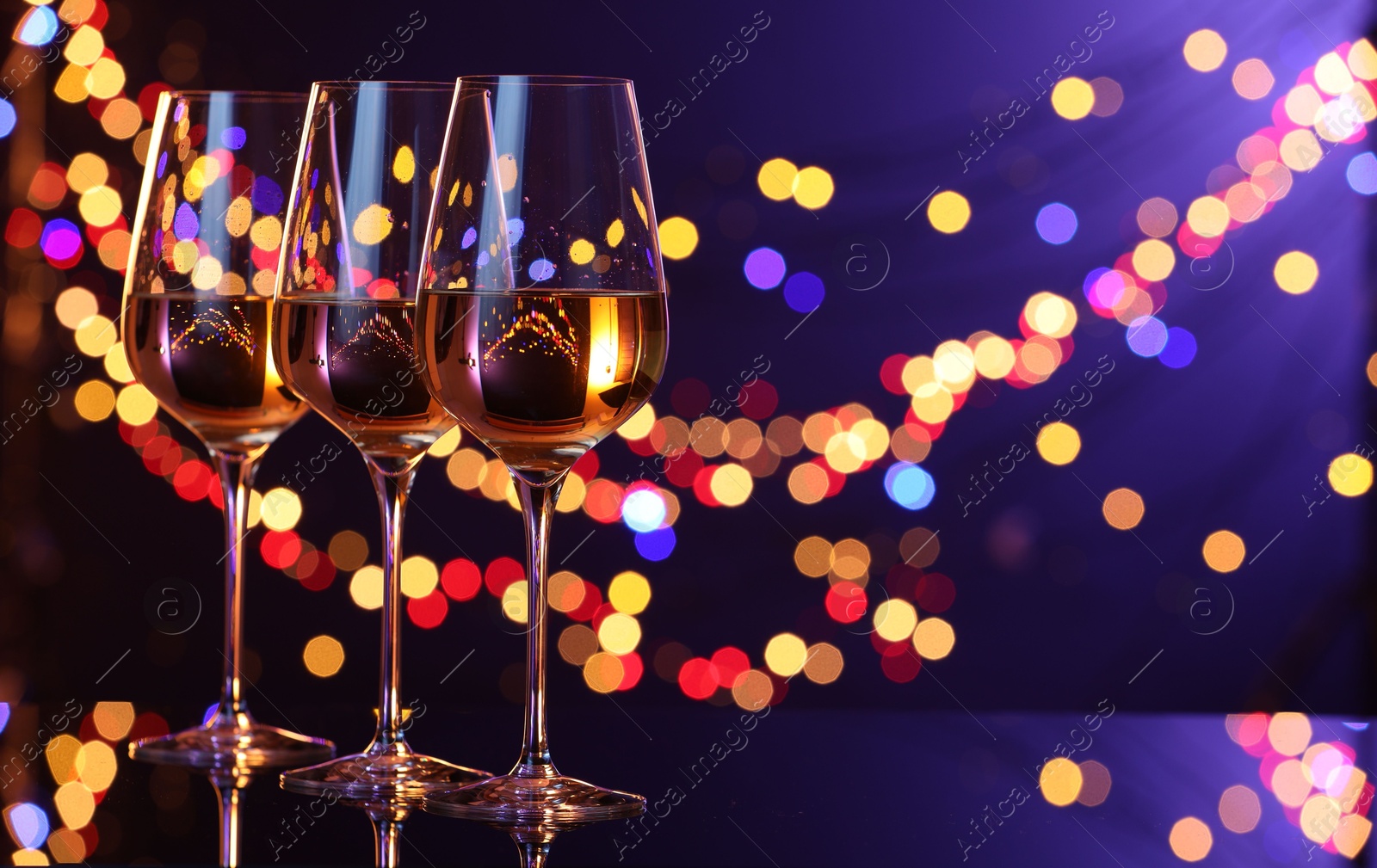 Photo of Tasty white wine in glasses on mirror surface against dark purple background with blurred lights, space for text. Bokeh effect