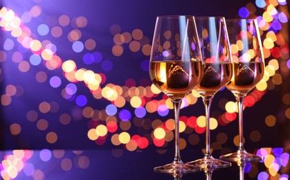 Photo of Tasty white wine in glasses on mirror surface against dark purple background with blurred lights, space for text. Bokeh effect