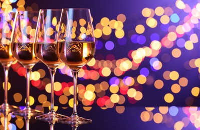 Photo of Tasty white wine in glasses on mirror surface against dark purple background with blurred lights, space for text. Bokeh effect