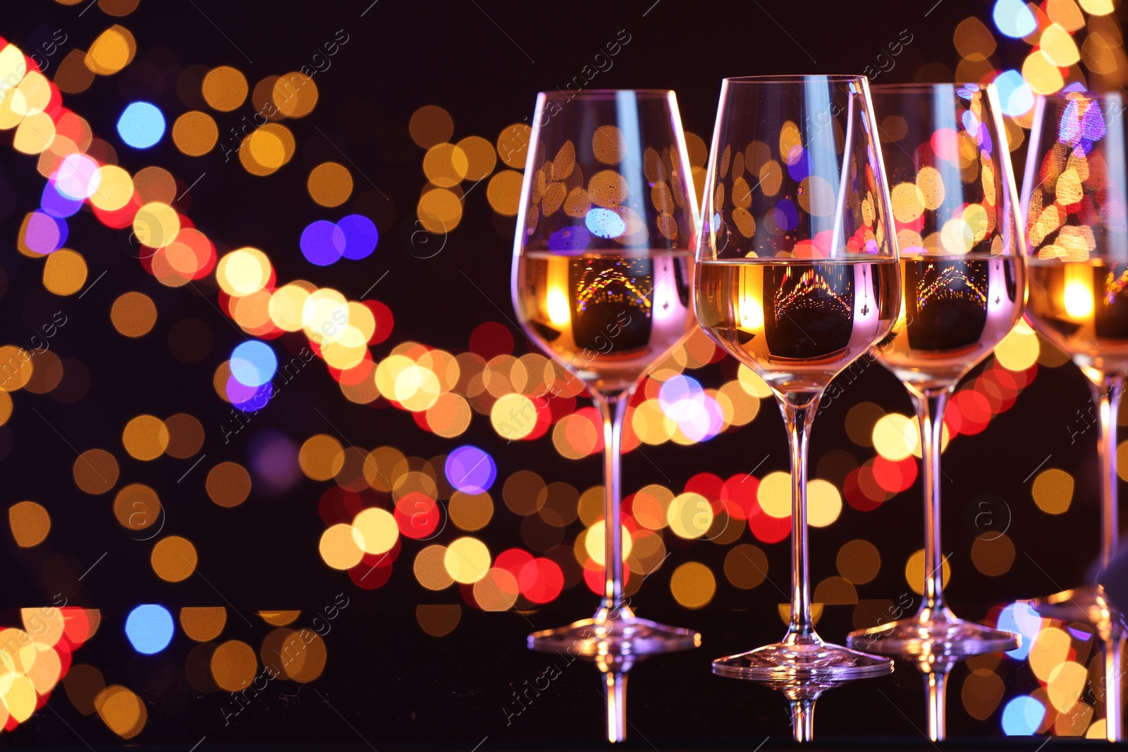 Photo of Tasty white wine in glasses on mirror surface against dark background with blurred lights, space for text. Bokeh effect