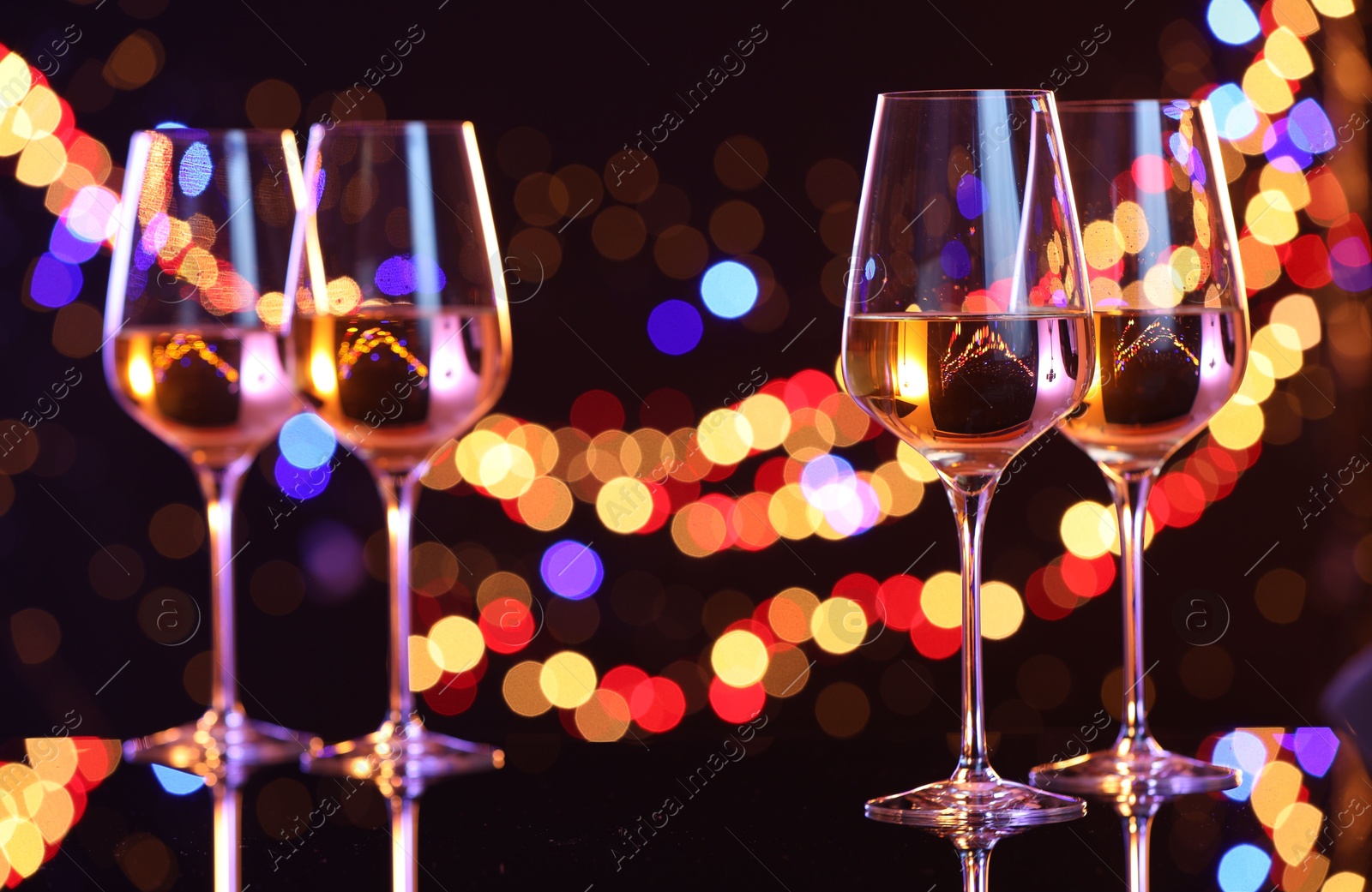 Photo of Tasty white wine in glasses on mirror surface against dark background with blurred lights, bokeh effect