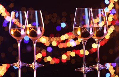 Tasty white wine in glasses on mirror surface against dark background with blurred lights, bokeh effect