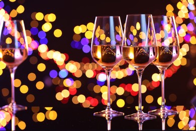 Photo of Tasty white wine in glasses on mirror surface against dark background with blurred lights, bokeh effect