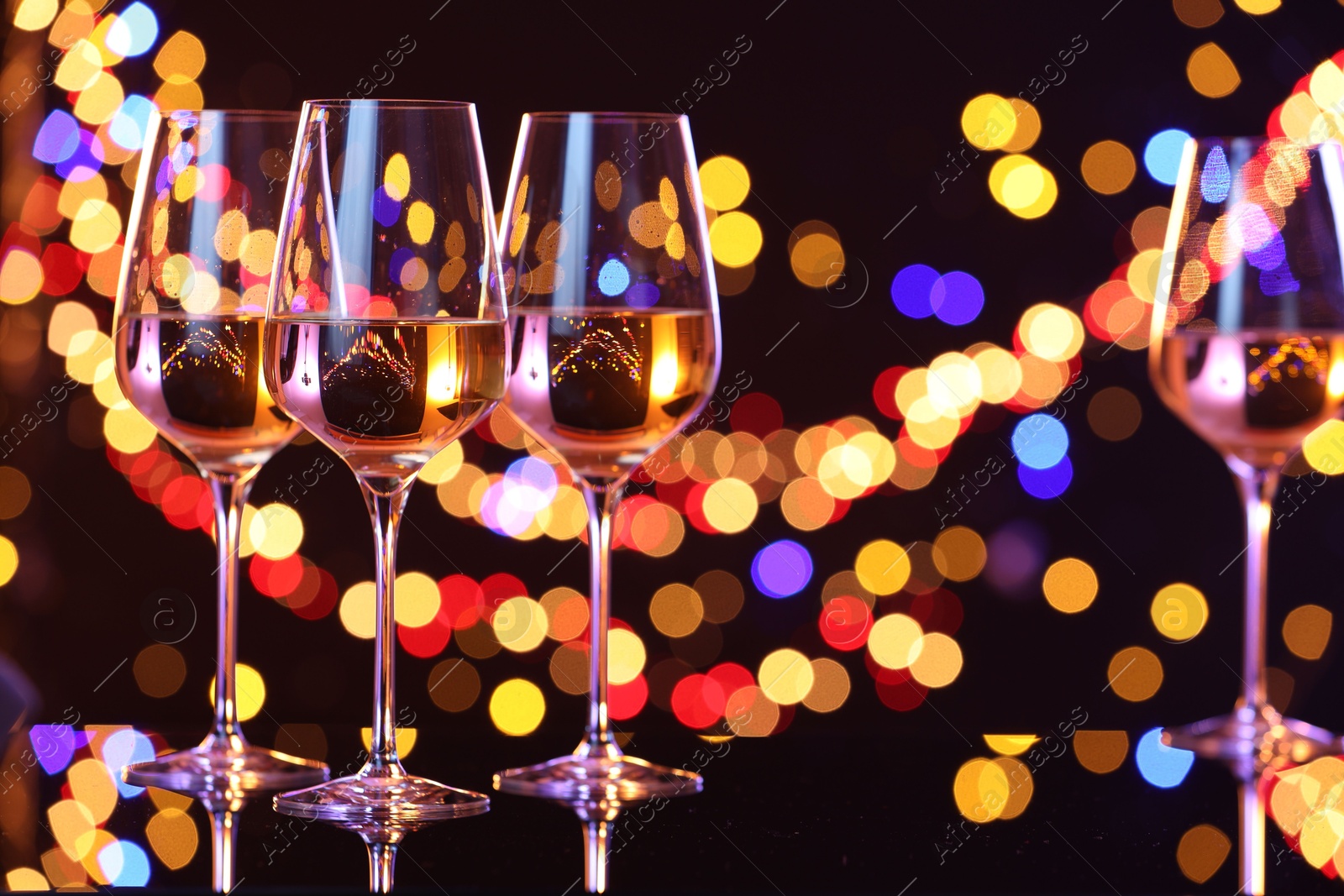 Photo of Tasty white wine in glasses on mirror surface against dark background with blurred lights, bokeh effect