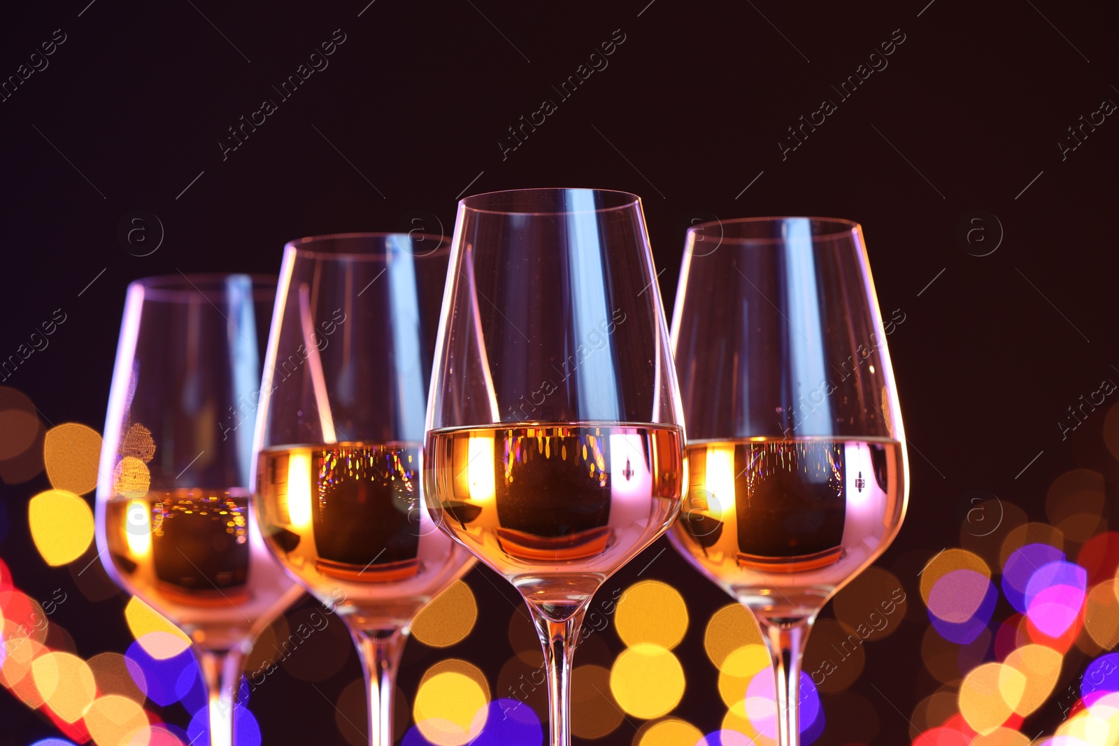 Photo of Tasty white wine in glasses against black background with blurred lights, closeup. Bokeh effect