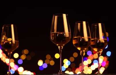 Photo of Tasty white wine in glasses against black background with blurred lights, low angle view. Bokeh effect