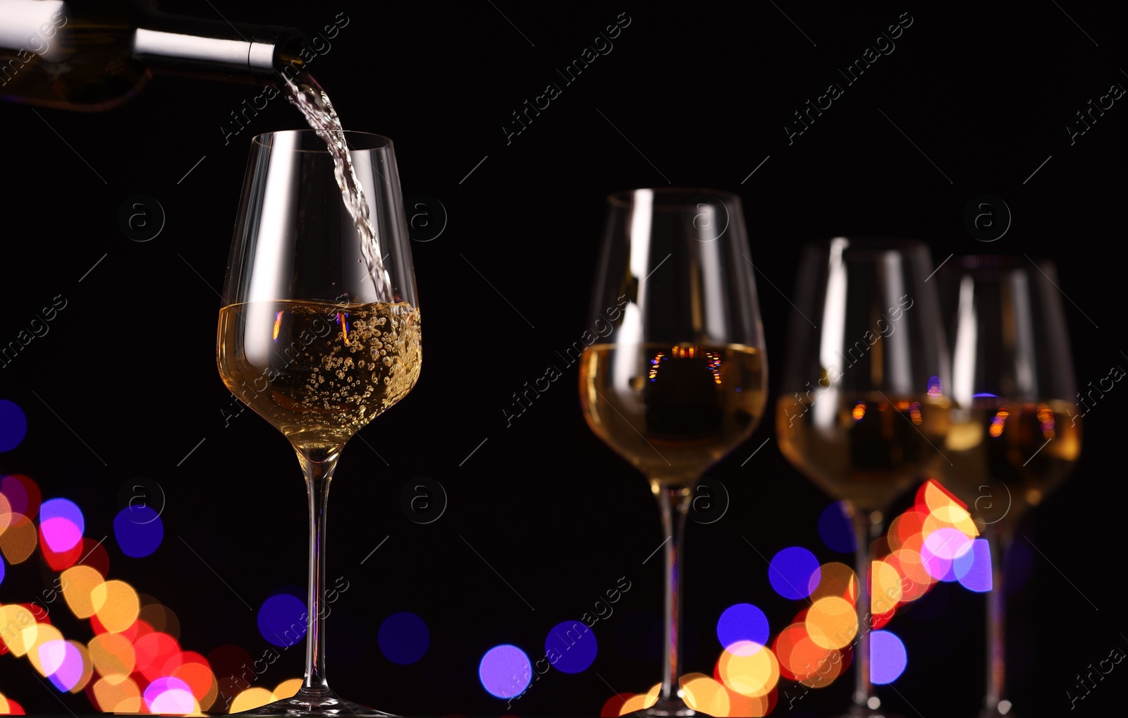 Photo of Pouring white wine into glass against black background with blurred lights, closeup. Bokeh effect