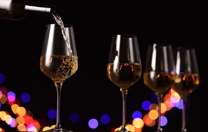 Photo of Pouring white wine into glass against black background with blurred lights, closeup. Bokeh effect