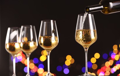 Photo of Pouring white wine into glass against black background with blurred lights, closeup. Bokeh effect