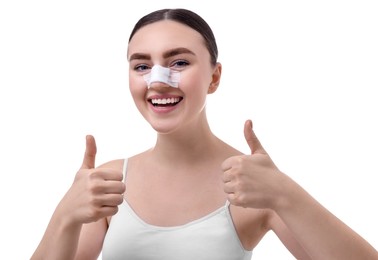 Photo of Woman with medical bandage on her nose after plastic surgery operation showing thumbs up against white background