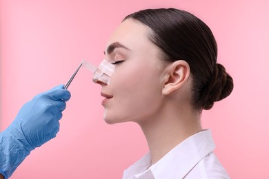 Photo of Doctor removing medical bandage from patient's nose after plastic surgery operation on pink background, closeup