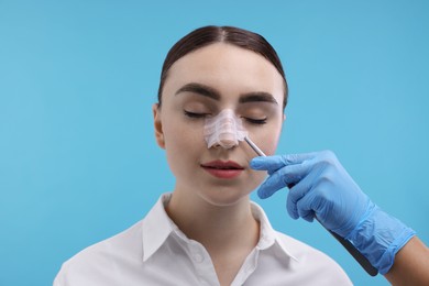 Doctor removing medical bandage from patient's nose after plastic surgery operation on light blue background, closeup