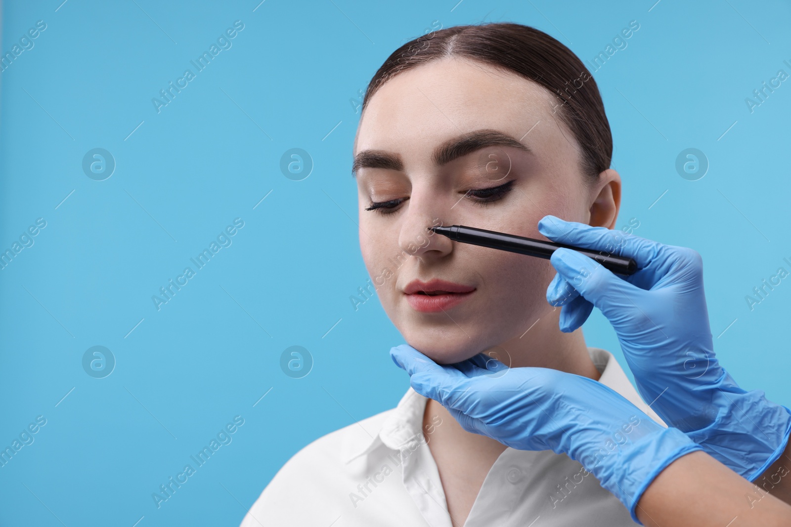 Photo of Doctor drawing marks on patient's nose for plastic surgery operation against light blue background, , closeup. Space for text