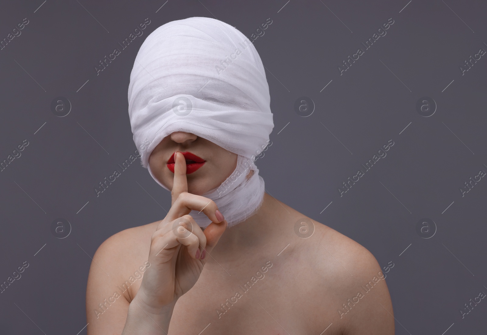 Photo of Woman with face wrapped in medical bandage after plastic surgery operation making silent gesture on grey background