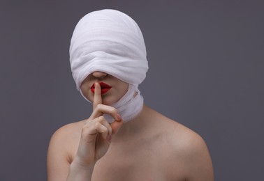 Photo of Woman with face wrapped in medical bandage after plastic surgery operation making silent gesture on grey background