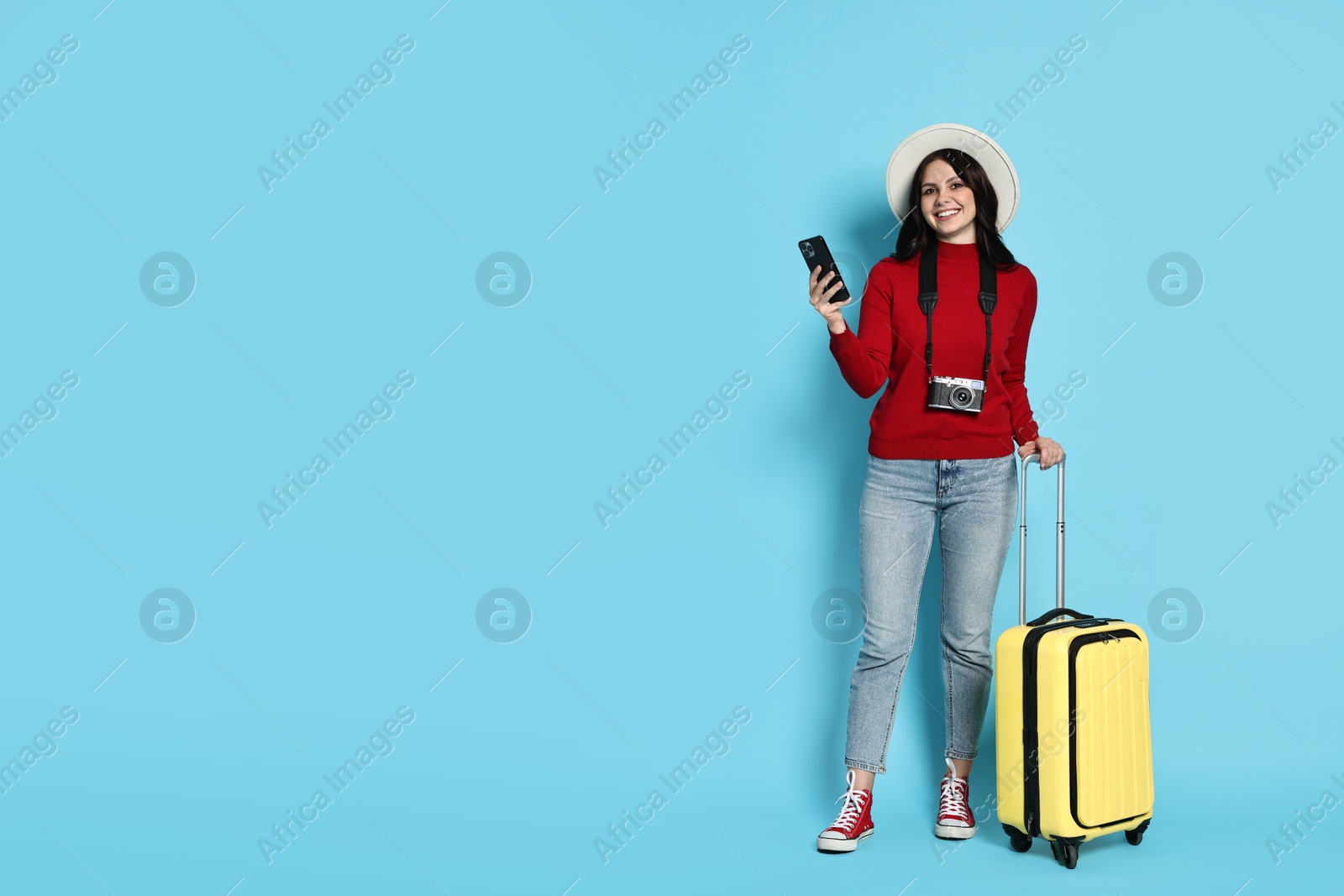 Photo of Young tourist in hat with camera, phone and suitcase on light blue background, space for text