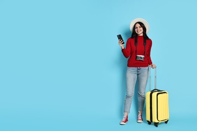 Young tourist in hat with camera, phone and suitcase on light blue background, space for text