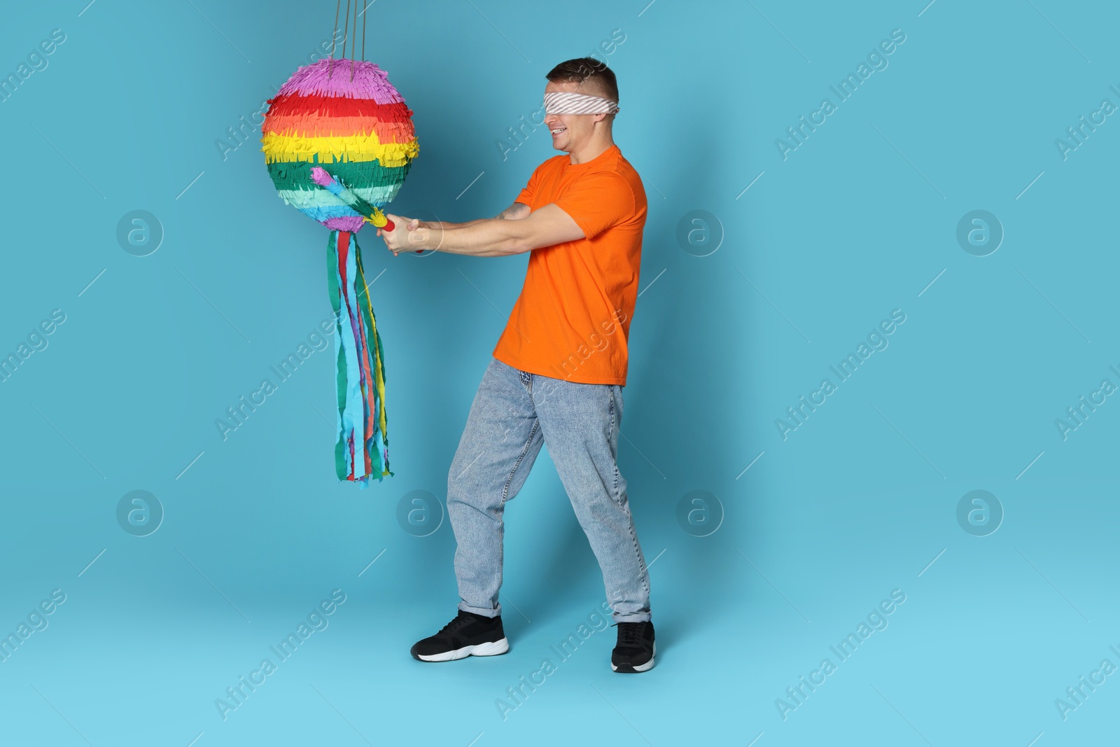 Photo of Man with tied eyes breaking pinata on light blue background