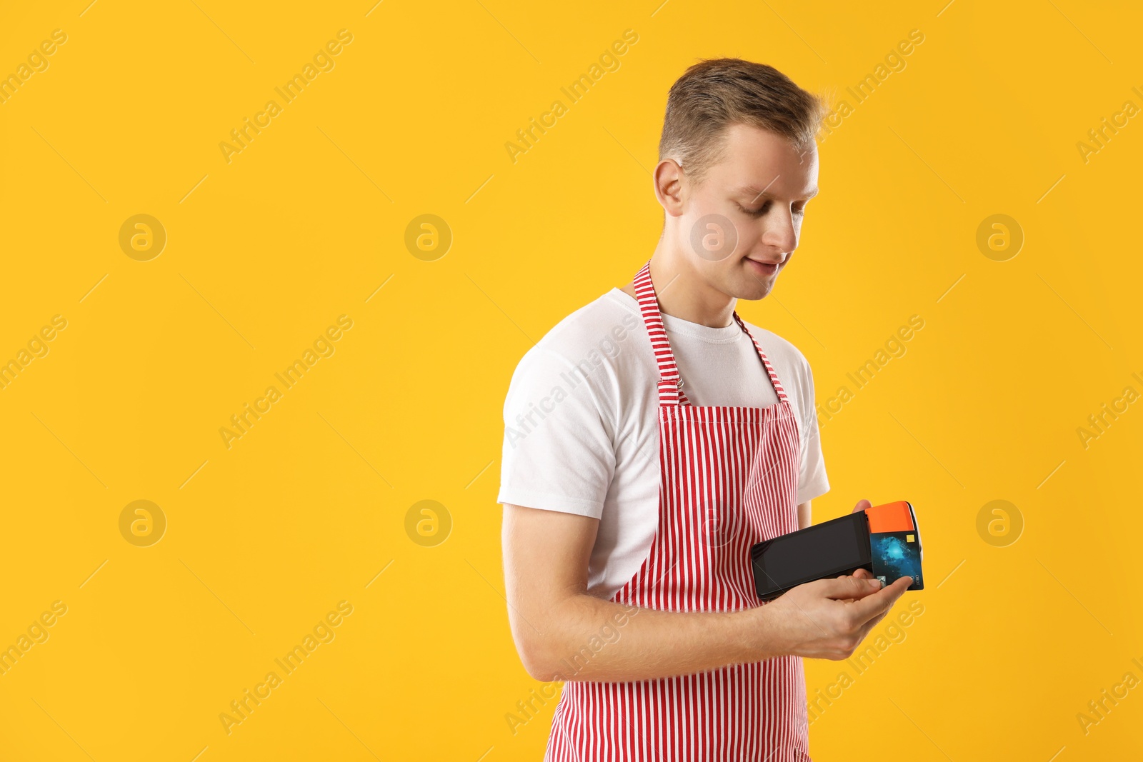 Photo of Young man in apron with payment terminal and debit card on yellow background