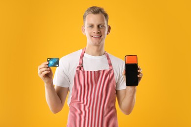 Happy young man in apron with payment terminal and debit card on yellow background