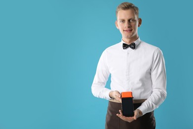 Photo of Happy waiter with payment terminal on light blue background, space for text