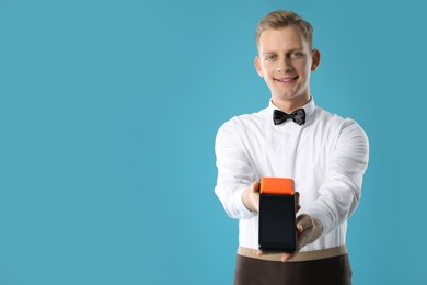 Photo of Happy waiter with payment terminal on light blue background, space for text