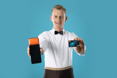 Photo of Happy waiter with payment terminal and debit card on light blue background