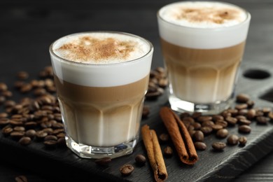 Photo of Tasty latte macchiato in glasses, coffee beans and cinnamon on black wooden table, closeup