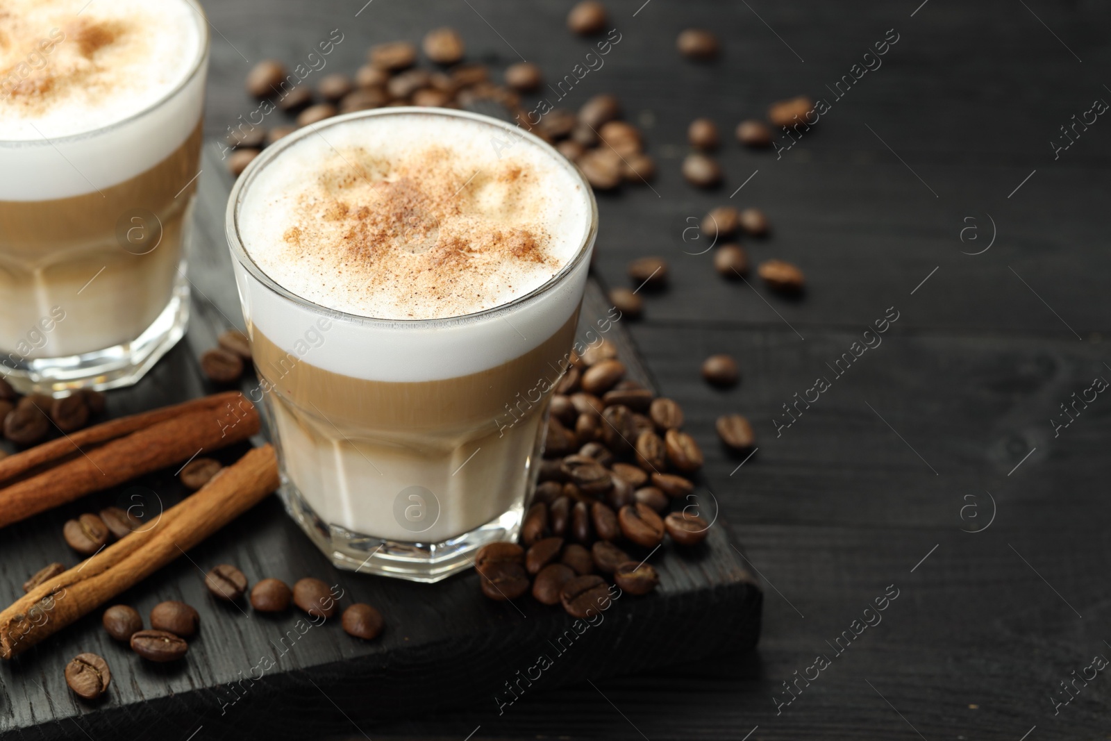 Photo of Tasty latte macchiato in glasses, coffee beans and cinnamon on black wooden table, closeup. Space for text