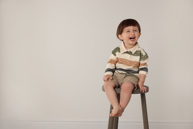 Photo of Emotional little boy sitting on stool against light grey background. Space for text