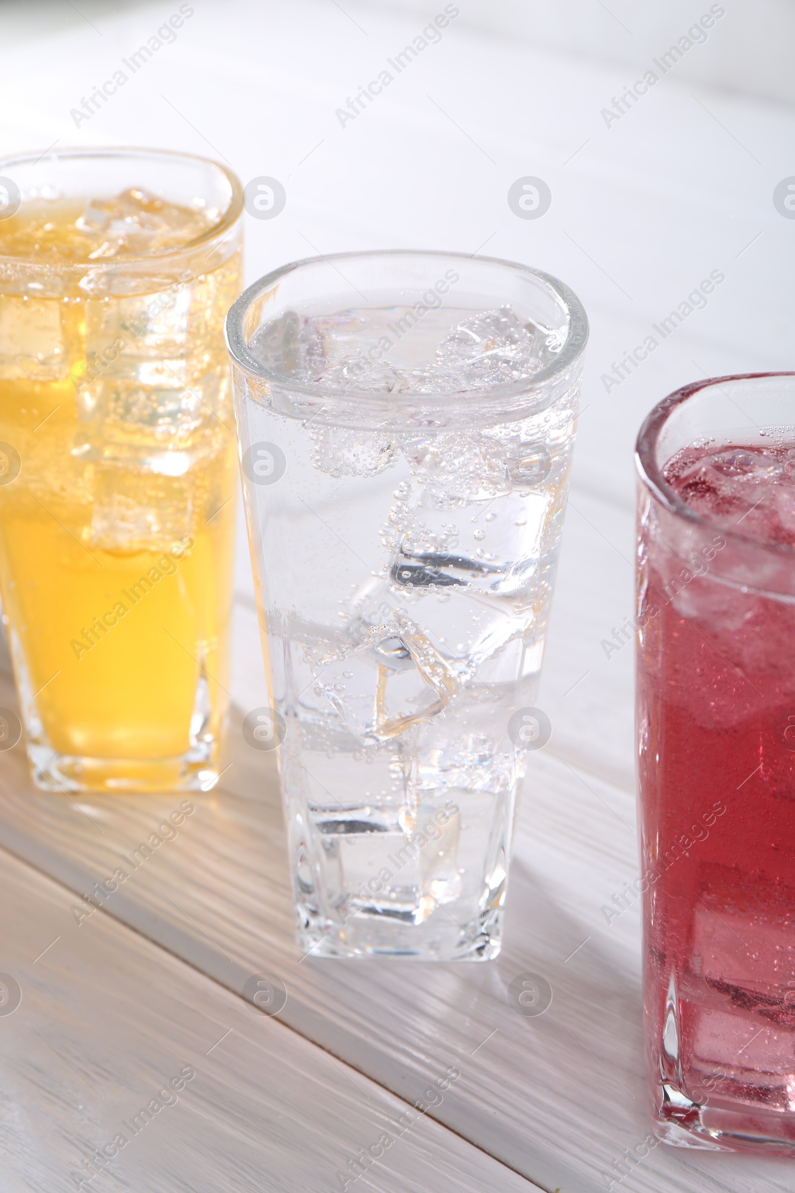 Photo of Soda water of different flavors with ice cubes in glasses on white wooden table, closeup