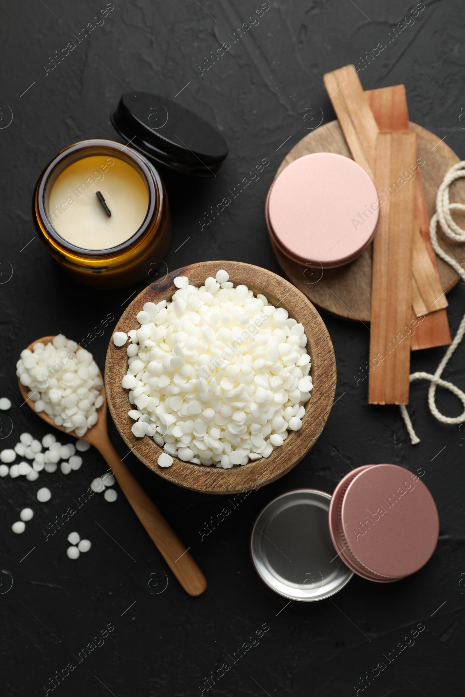 Photo of Soy wax in bowl, candle, wooden wicks, twine and jars on black table, flat lay