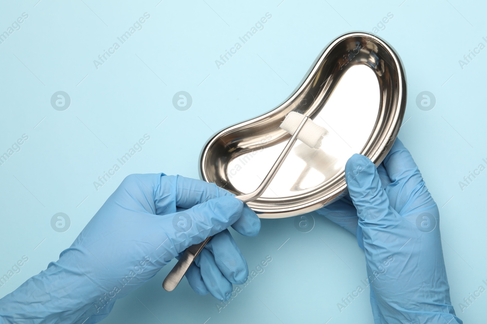 Photo of Doctor holding surgical tweezers with gauze and tray on light blue background, top view