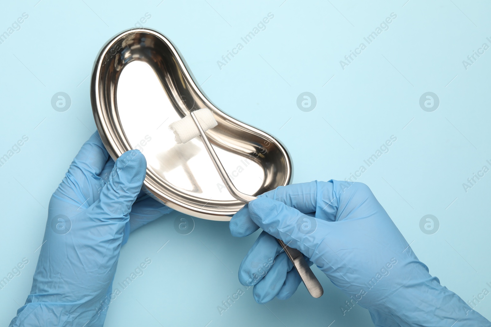 Photo of Doctor holding surgical tweezers with gauze and tray on light blue background, top view