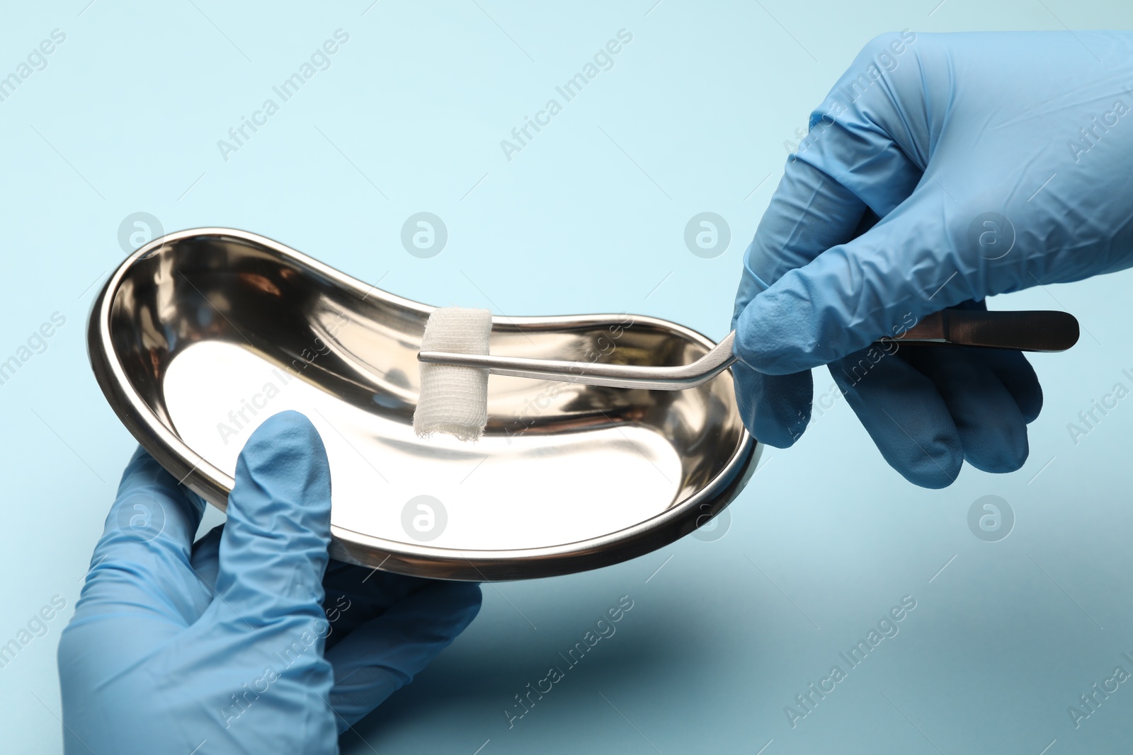 Photo of Doctor holding surgical tweezers with gauze and tray on light blue background, closeup