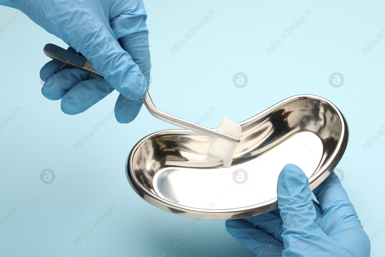 Photo of Doctor holding surgical tweezers with gauze and tray on light blue background, closeup