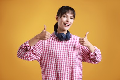 Photo of Happy woman with headphones showing thumbs up on orange background. Like gesture