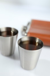 Photo of Hip flask and cups with whiskey on white table, closeup