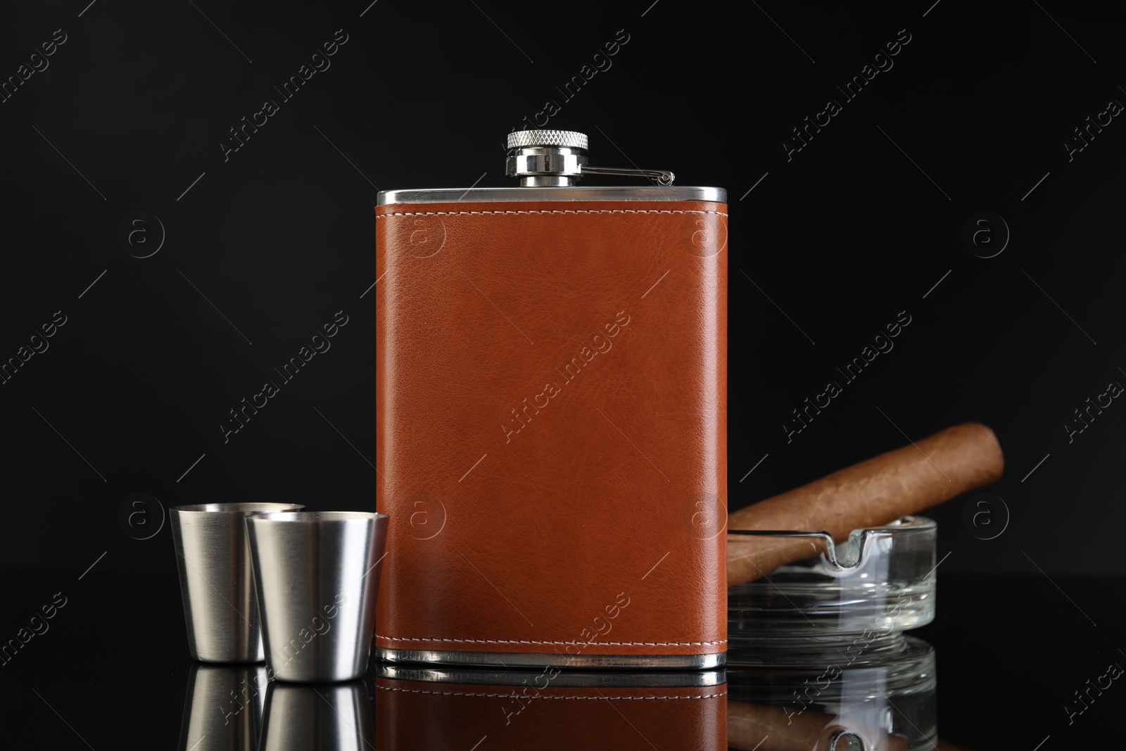 Photo of Hip flask, cups, cigar and ashtray on black mirror surface