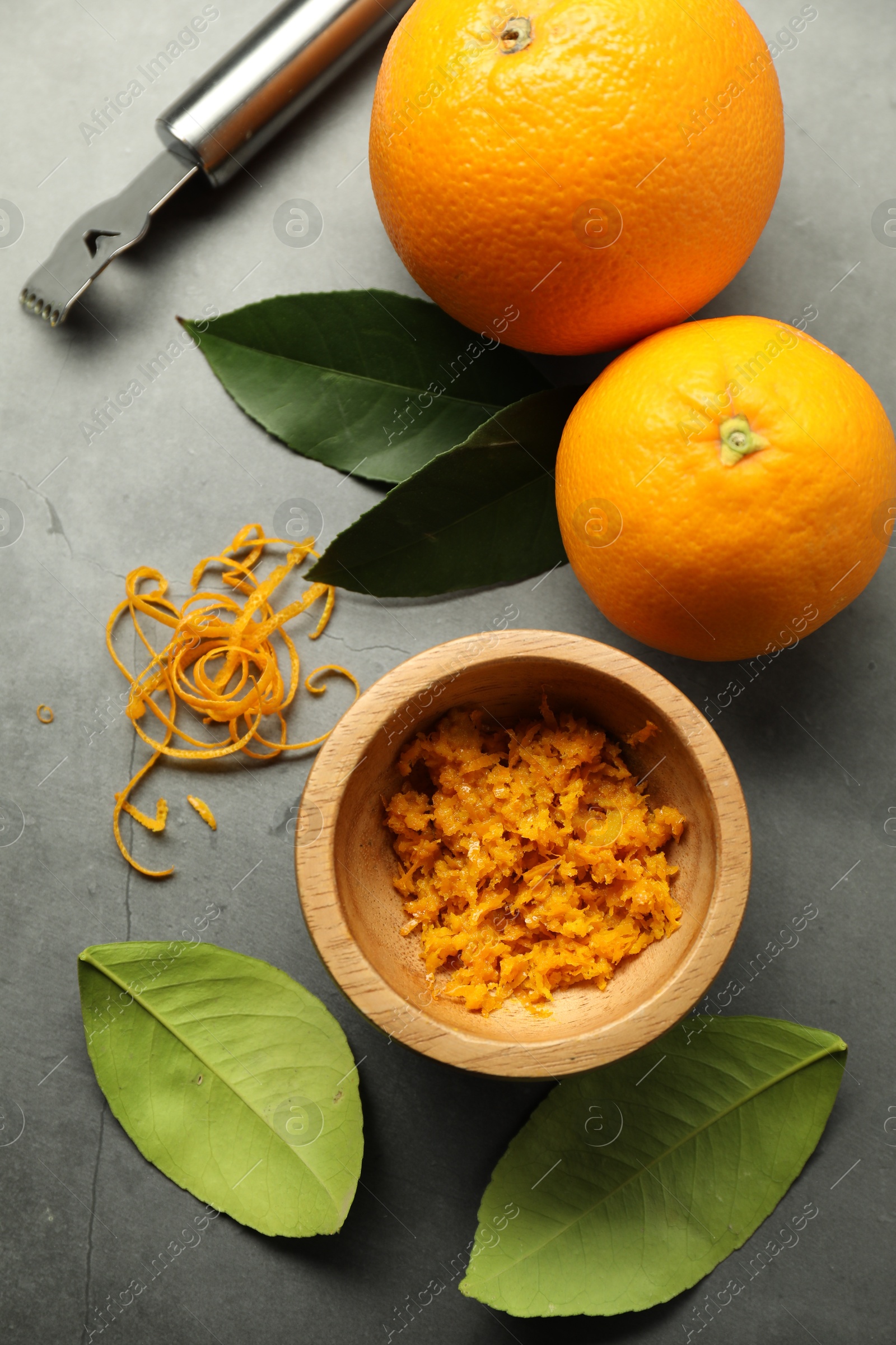 Photo of Orange zest, zester tool and fresh fruits on gray textured table, flat lay