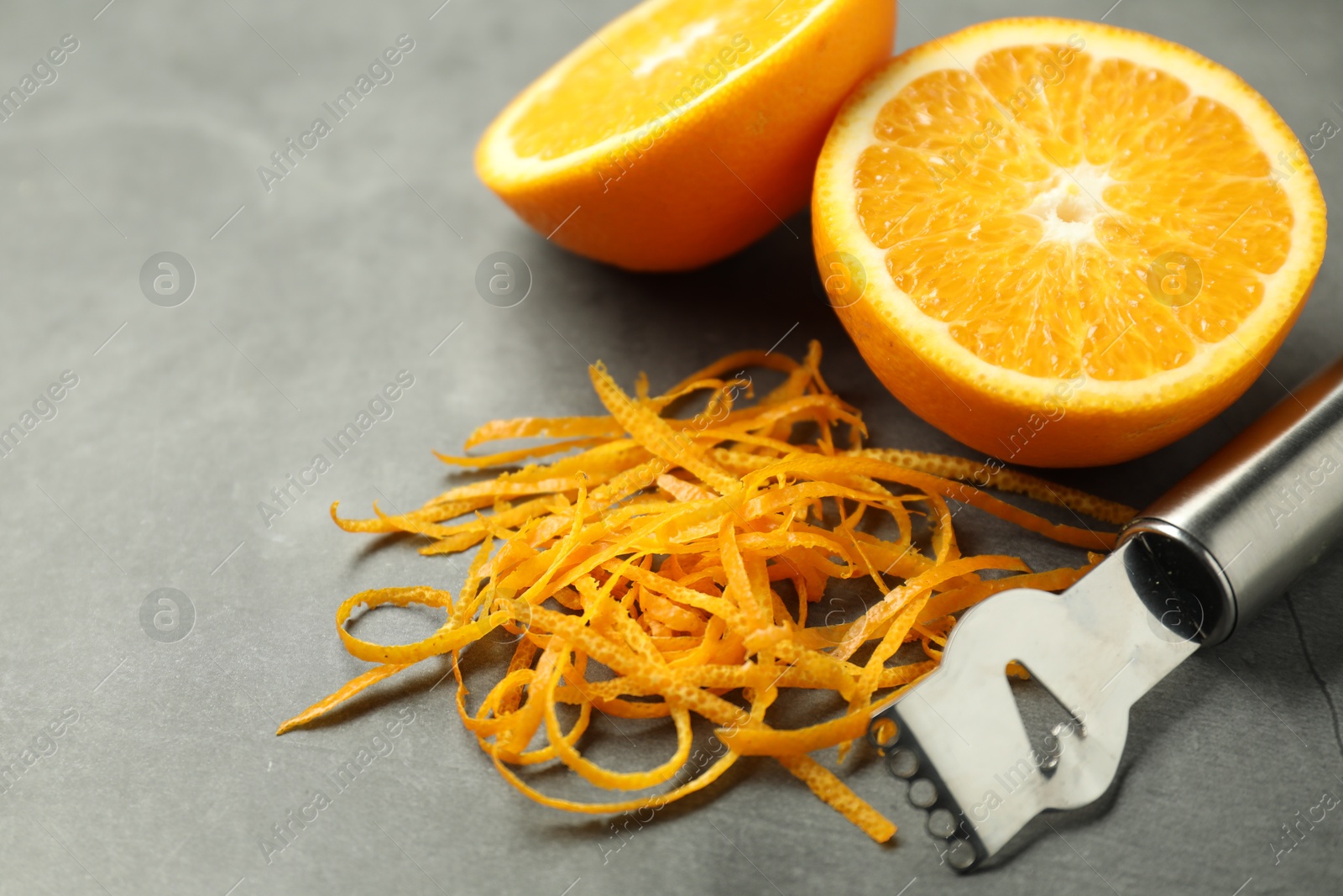 Photo of Orange zest, zester tool and fresh fruit pieces on gray textured table, closeup. Space for text