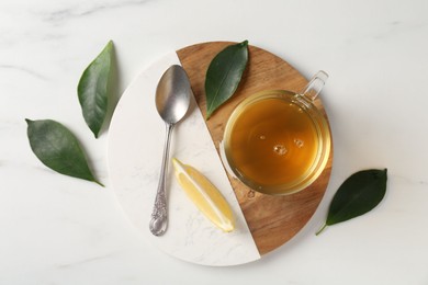 Photo of Refreshing green tea in cup, spoon, leaves and slice of lemon on white marble table, flat lay