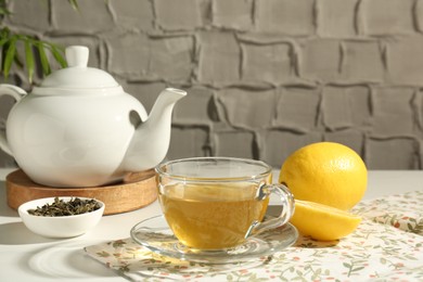 Refreshing green tea in cup, lemon and dry leaves on white table against grey textured wall, closeup