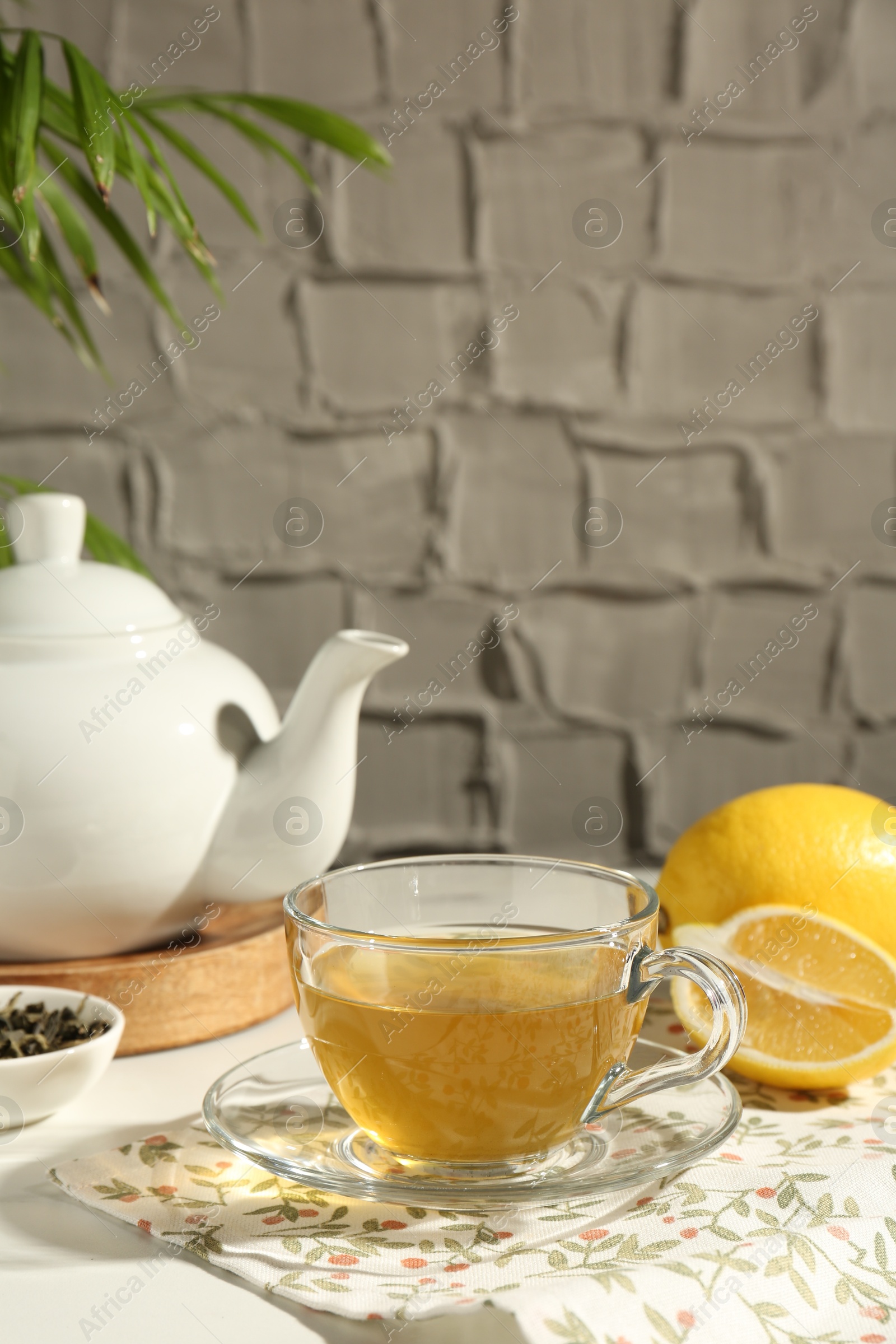 Photo of Refreshing green tea in cup, lemon and dry leaves on white table against grey textured wall