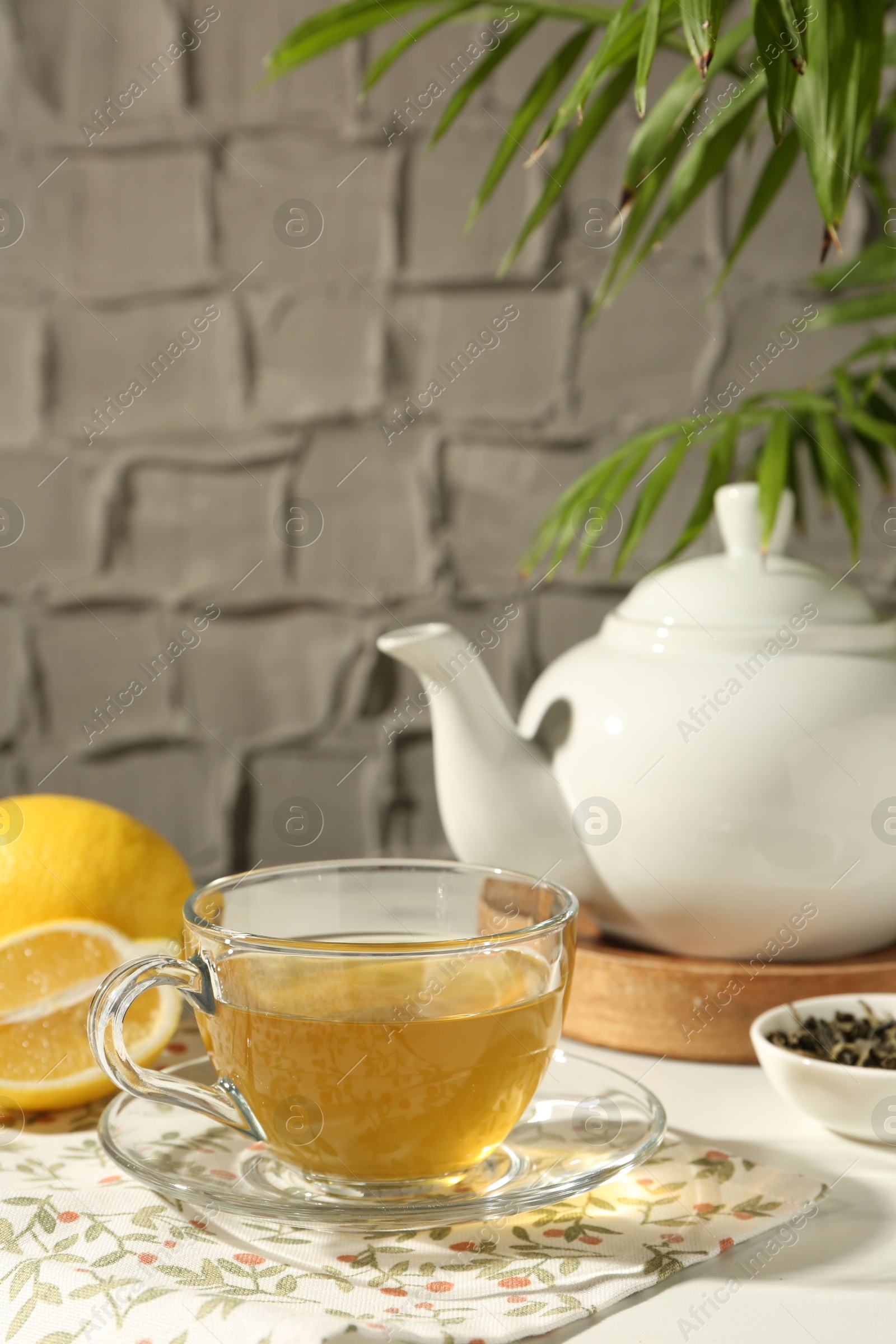 Photo of Refreshing green tea in cup, lemon and dry leaves on white table against grey textured wall