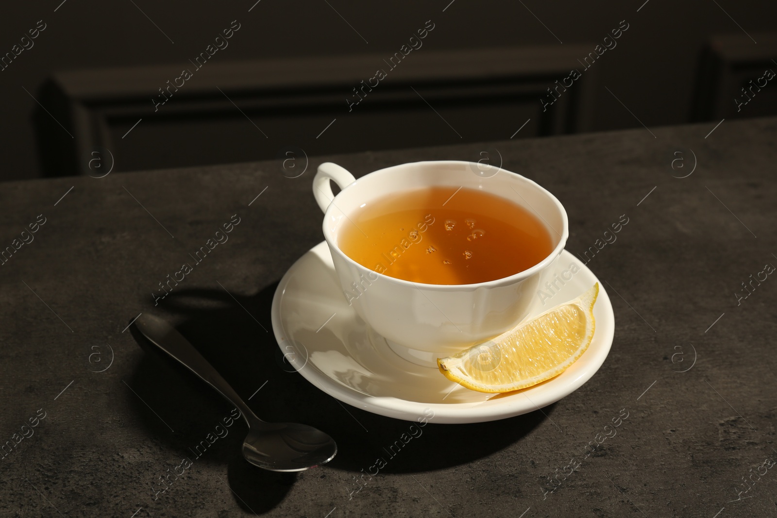 Photo of Refreshing green tea in cup, slice of lemon and spoon on dark textured table