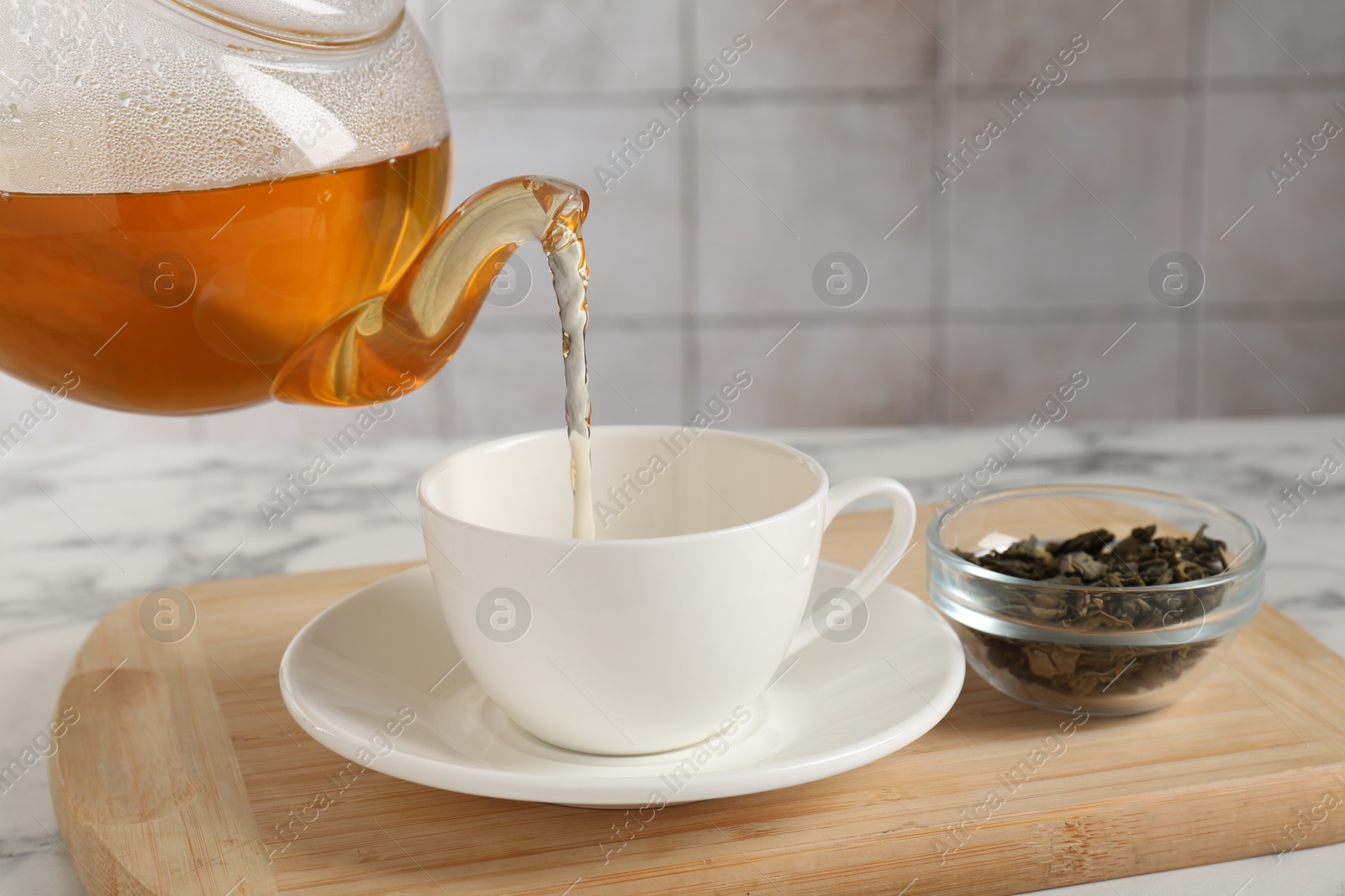 Photo of Pouring refreshing green tea from teapot into cup at white marble table, closeup