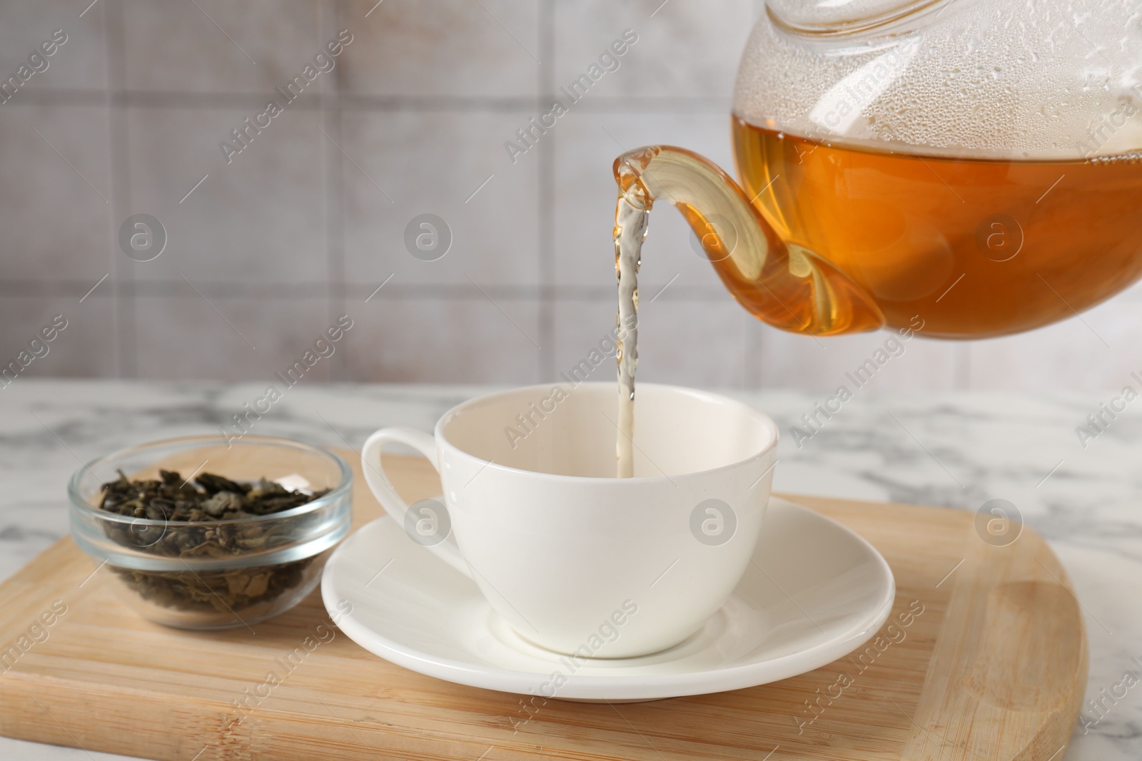 Photo of Pouring refreshing green tea from teapot into cup at white marble table, closeup