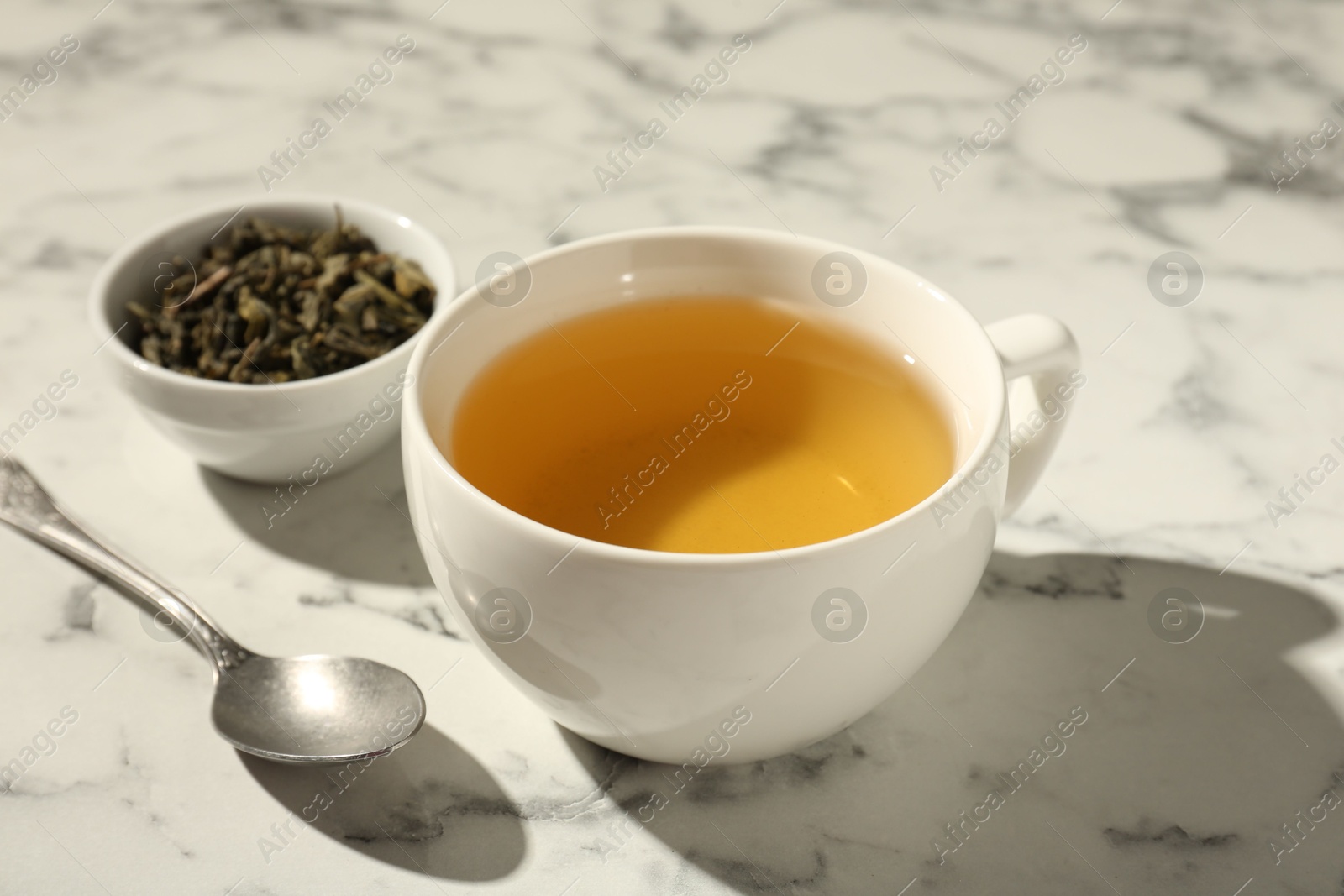 Photo of Refreshing green tea in cup, dry leaves and spoon on white marble table, closeup