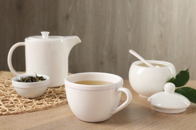 Photo of Refreshing green tea in cup, sugar bowl, leaves and teapot on wooden table